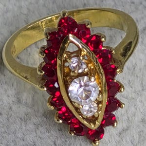 May include: A gold ring with a central setting of clear stones, surrounded by a cluster of small, red gemstones. The ring's band is plain gold. The central setting is elongated.