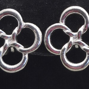 May include: A pair of silver-colored stud earrings. Each earring features a four-leaf clover design, with interlocking circular shapes. The earrings are displayed against a black background.