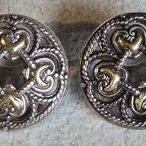 May include: A pair of silver-toned stud earrings with gold-colored accents. The earrings have a circular design with an open filigree pattern and a decorative rope-like border. The gold accents are in the center and at the four points of the design.