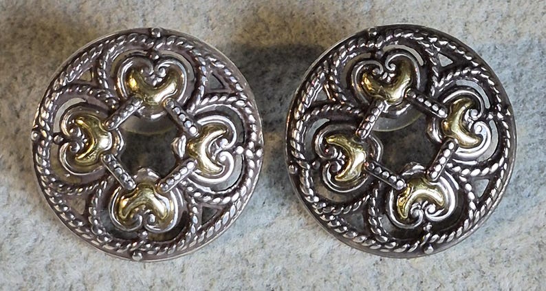 May include: A pair of silver-toned stud earrings with gold-colored accents. The earrings have a circular design with an open filigree pattern and a decorative rope-like border. The gold accents are in the center and at the four points of the design.