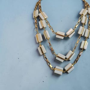 May include: A multi-strand necklace featuring gold-tone metal chains interspersed with rectangular, off-white, smooth-surfaced beads. The design consists of three cascading layers of linked chains, each adorned with these uniform, light-colored rectangular accents.