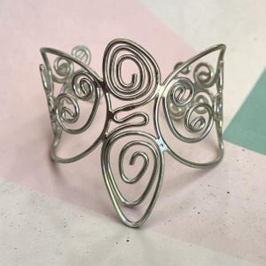 May include: A silver-toned metal cuff bracelet featuring an openwork design composed of multiple interconnected wire spirals and loops. The structure is symmetrical, with a prominent central teardrop-shaped spiral and smaller circular swirls.