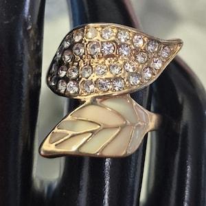 May include: A gold-toned ring featuring a leaf design. The top leaf is encrusted with clear rhinestones, while the lower leaf has a cream and light brown enamel inlay. The ring is set against a dark background.