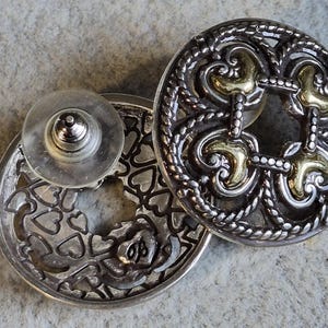 May include: Silver-toned stud earrings with intricate filigree and gold-colored accents. One earring features a heart-shaped design, while the other has a decorative openwork pattern. The earrings have a post and backing for secure wear.