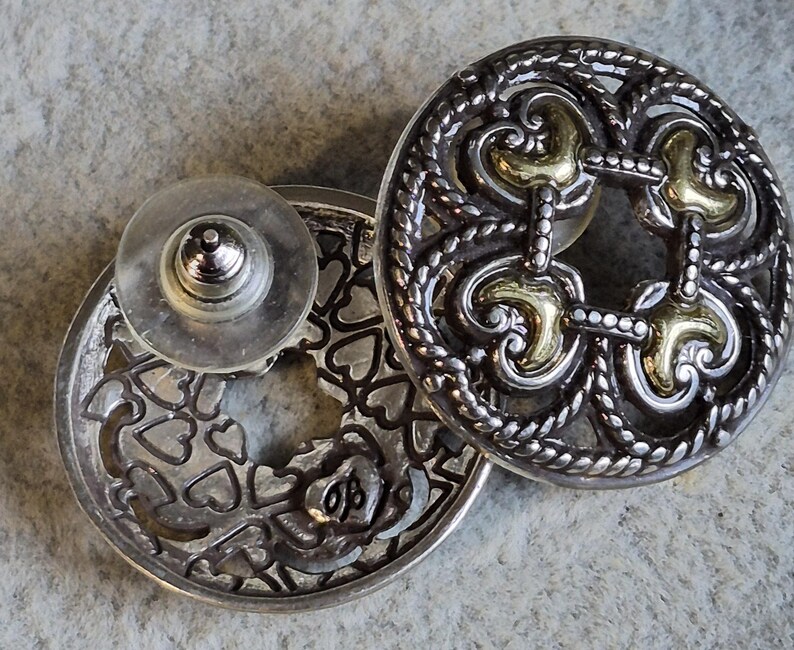 May include: Silver-toned stud earrings with intricate filigree and gold-colored accents. One earring features a heart-shaped design, while the other has a decorative openwork pattern. The earrings have a post and backing for secure wear.