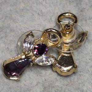 May include: Gold-toned stud earrings with a cluster of gemstones. The earrings feature a teardrop-shaped purple gem, a round purple gem, and two clear, marquise-cut gems. The earring posts have clear plastic backings.