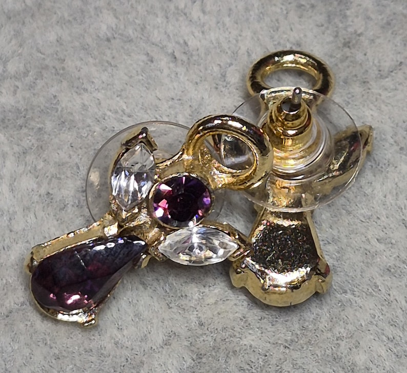 May include: Gold-toned stud earrings with a cluster of gemstones. The earrings feature a teardrop-shaped purple gem, a round purple gem, and two clear, marquise-cut gems. The earring posts have clear plastic backings.