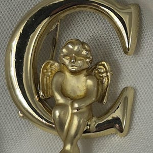 May include: A gold-toned brooch featuring a seated cherub figure within a stylized letter "C". The cherub has detailed wings and is positioned inside the curve of the letter. The brooch has a polished finish.