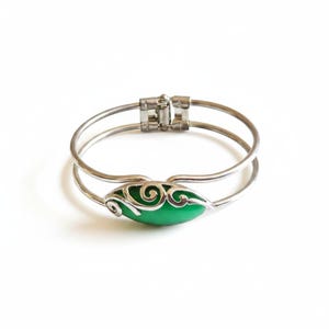 May include: A silver-toned metal hinged bangle bracelet featuring a double-band design. The center focal point is an oval-shaped, smooth green stone inlay accented with decorative silver-toned metal swirls that wrap over the top of the stone.