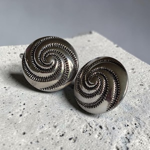 May include: A pair of circular silver-toned metal buttons featuring a raised, spiraling pattern that radiates from the center. The surface of each button is polished and reflective, with textured, notched detailing along the spiral grooves.