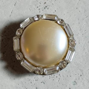 May include: A round button featuring a large, smooth, cream-colored faux pearl center. The pearl is surrounded by a circular border of alternating clear baguette-cut and round-cut rhinestones set in a metallic frame, creating a decorative edge.