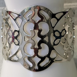 May include: A silver-colored cuff bracelet with an openwork design. The bracelet features a repeating pattern of curved shapes and circles. The metal cuff is wide and wraps around the wrist. The bracelet is shown on a white surface.