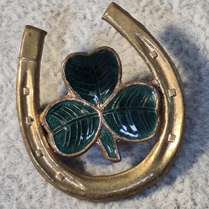 May include: A gold-colored horseshoe pin with a green shamrock in the center. The shamrock has detailed green enamel leaves. The horseshoe has small square indentations along the edges.