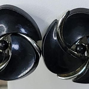 May include: A pair of vintage black clip-on earrings. Each earring features a swirling, three-petal design with silver-tone metal accents. A small, black bead is centered in each earring. The earrings are likely from the mid-century era.