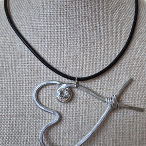 May include: A silver-toned metal pendant necklace with a black cord. The pendant features a heart-shaped design with a clear, round gemstone and a wrapped section. The necklace is displayed on a neutral-toned fabric.