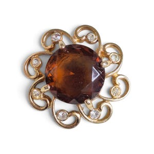 May include: A gold-toned brooch featuring a large, round, faceted amber-colored central stone. The stone is surrounded by a decorative metal frame with swirling filigree patterns and small, clear, round inset stones at each curve of the design.