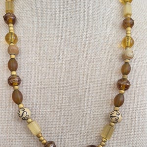 May include: A beaded necklace featuring a variety of amber and brown-toned beads. The necklace includes round, oval, and cylindrical beads, with some adorned with decorative patterns. The beads are strung together with gold-colored spacers.