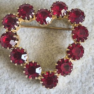 May include: A vintage heart-shaped brooch with a gold-tone setting. The brooch is adorned with numerous round, faceted, deep red gemstones. The gemstones are set closely together, forming the heart shape. A pin is attached to the back.