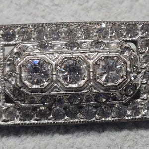 May include: A rectangular, silver-toned brooch with a central design of three large, clear, faceted stones. The brooch is encrusted with numerous smaller, clear stones, creating a sparkling effect. The design is intricate and detailed.
