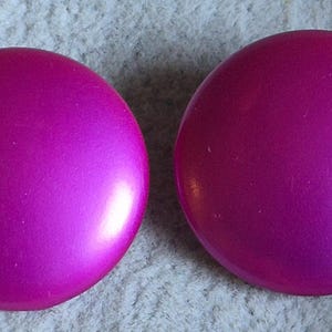 May include: Two round, magenta-colored buttons with a glossy finish. The buttons are smooth and appear to be made of plastic or a similar material. They are set against a neutral, textured background.