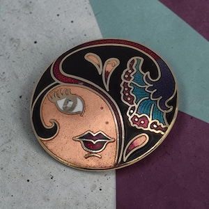 May include: A round metal brooch featuring a stylized face design with a copper-toned complexion, black hair, and a dark red lip. The design includes a decorative floral element in shades of teal, purple, and magenta, all outlined in gold-tone metal.