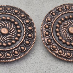 May include: Two antique copper-colored circular decorative elements. Each has a central spiral design, surrounded by a ring of small beads, a rope-like border, and an outer ring of larger beads. The overall design is symmetrical and ornate.