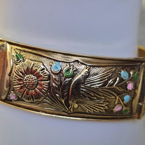 May include: Gold-toned hinged bangle bracelet with embossed floral and bird design. The bracelet features a detailed relief of a bird, flowers, and leaves, with accents of pink, blue, and green enamel.