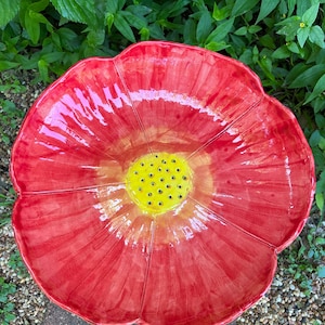 May include: A large, red ceramic flower-shaped bird bath. The petals are a vibrant red, and the center is yellow with small black dots. The bird bath is placed outdoors, surrounded by green foliage and small pebbles.