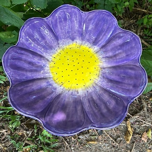 May include: A large, purple ceramic flower-shaped bird bath with a yellow center dotted with black spots. The bird bath is sitting on the ground surrounded by green foliage.