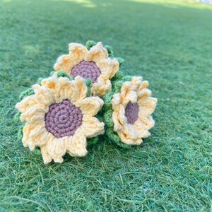 May include: Three crocheted sunflowers in yellow and green, arranged on a green grassy surface.