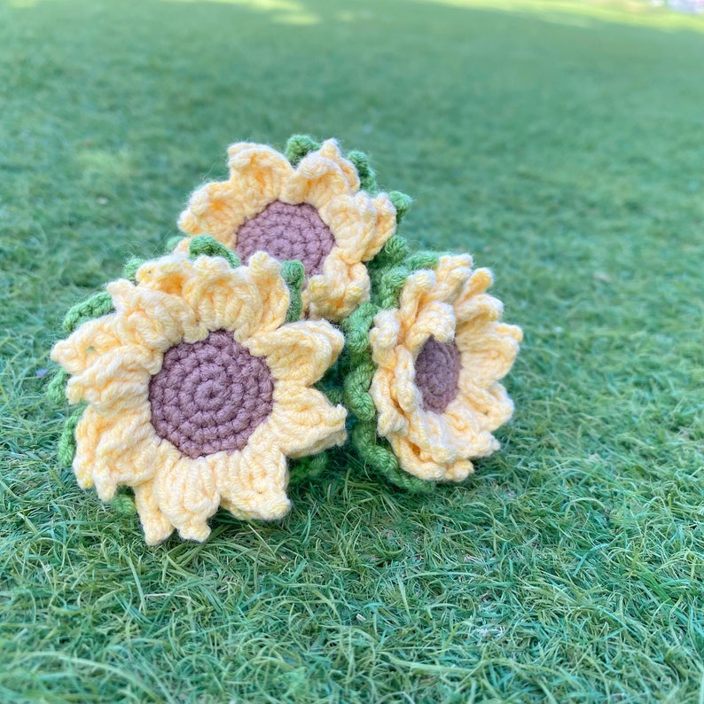 May include: Three crocheted sunflowers in yellow and green, arranged on a green grassy surface.