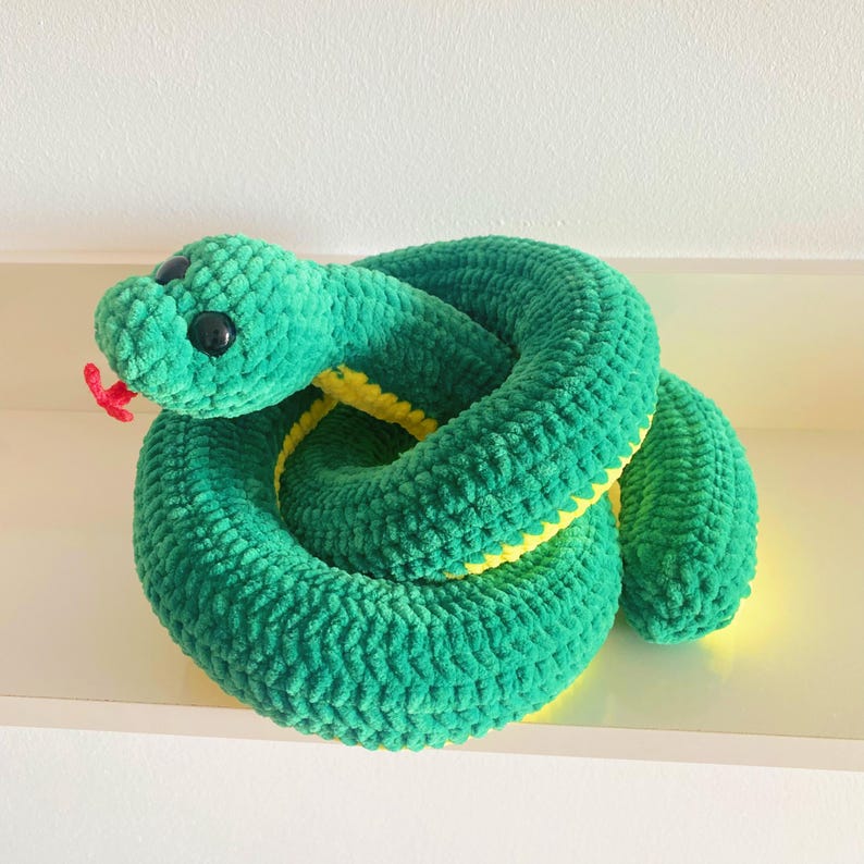 May include: A green crocheted snake plush toy with yellow accents and black eyes. The snake is curled up in a coil.