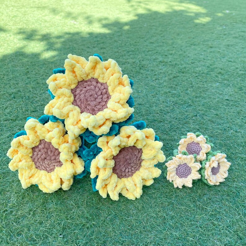 May include: Four crocheted sunflowers in various sizes, with yellow petals and brown centers. The sunflowers are arranged in a cluster on a green grassy surface.