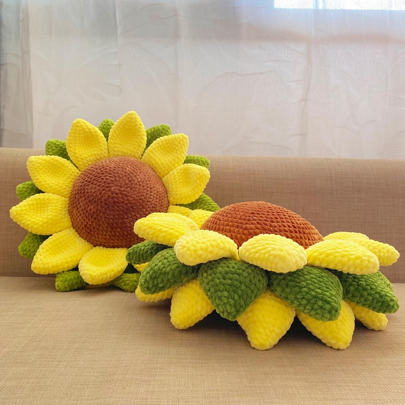 May include: Two handmade sunflower-shaped pillows. The pillows feature a plush, textured design with yellow petals, green leaves, and a brown center. The pillows are placed on a neutral-toned surface, showcasing their vibrant colors and unique shape.
