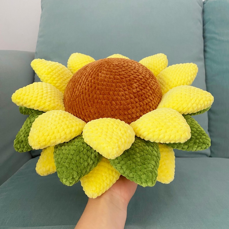 May include: A handmade, plush sunflower pillow with a brown center, yellow petals, and green leaves. The soft, textured pillow is designed to resemble a sunflower and is likely used for home decor.