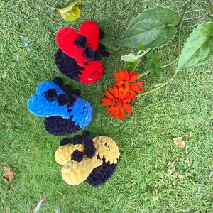 May include: Three handmade crochet ladybug toys in red, blue, and yellow with black accents, resting on green grass. The ladybugs have a rounded shape with a textured, plush appearance. Orange flowers and green leaves are also visible.
