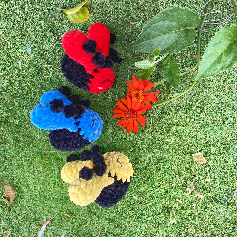 May include: Three handmade crochet ladybug toys in red, blue, and yellow with black accents, resting on green grass. The ladybugs have a rounded shape with a textured, plush appearance. Orange flowers and green leaves are also visible.