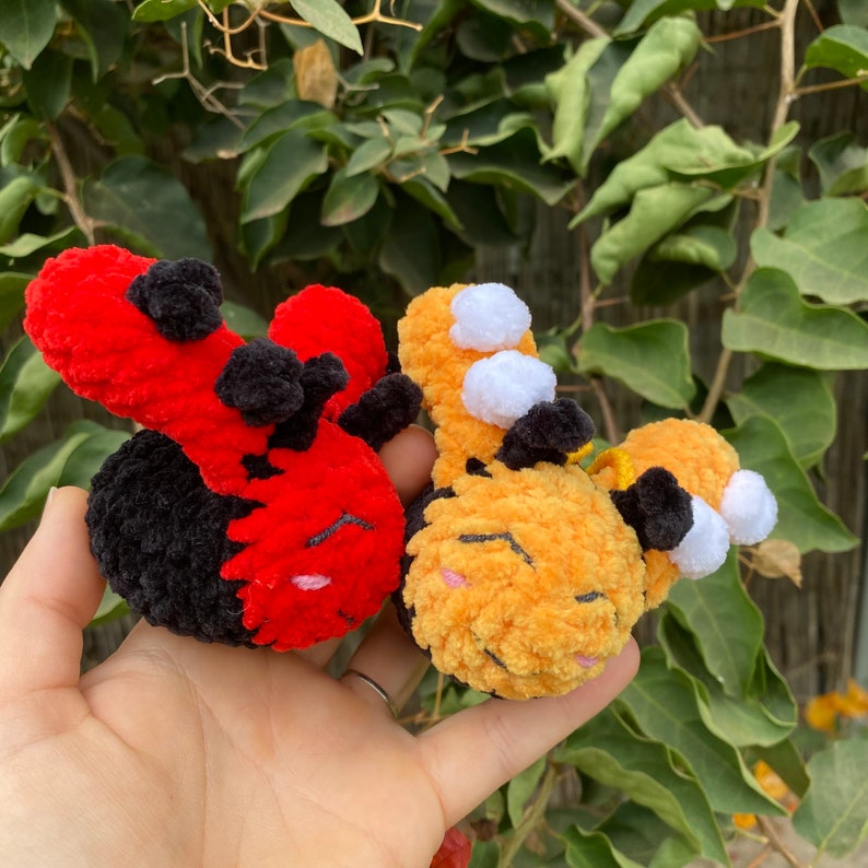May include: Two handmade crochet plush toys: a red and black ladybug and a yellow and black bumblebee. The ladybug has black spots and the bumblebee has white accents. Both have closed eyes and are held in a hand.