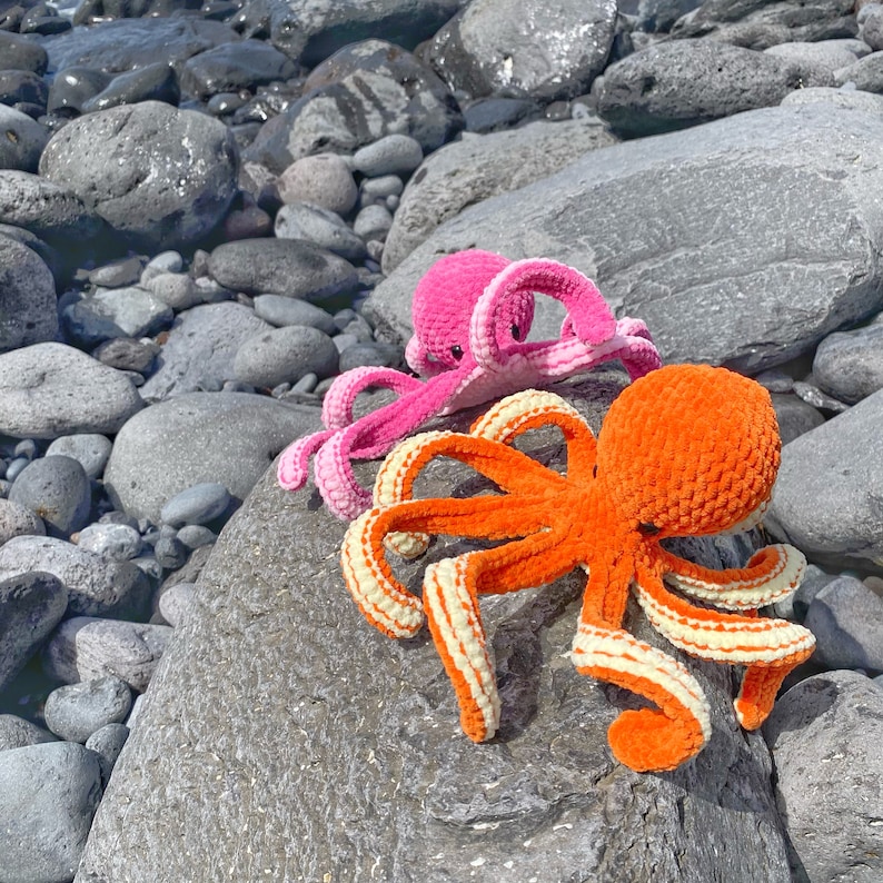 以下が含まれることがあります： グレーの岩の背景に、ピンクとオレンジの2つの編みぐるみタコ。