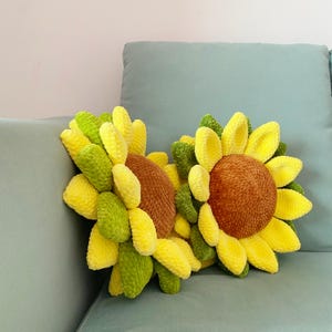 May include: Two handmade sunflower pillows with yellow, green, and brown details. The pillows are made of a soft, textured material and are designed to resemble sunflowers. The pillows are placed on a light blue couch.