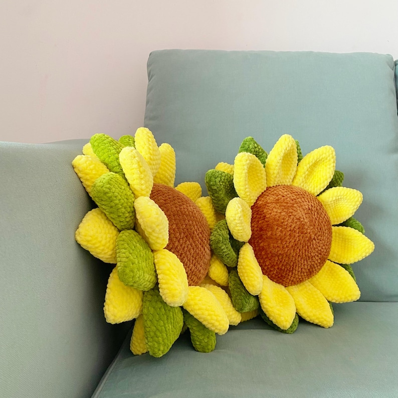 May include: Two handmade sunflower pillows with yellow, green, and brown details. The pillows are made of a soft, textured material and are designed to resemble sunflowers. The pillows are placed on a light blue couch.