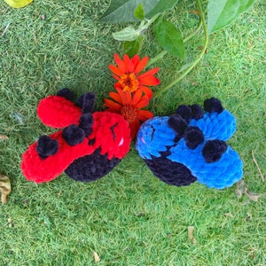 May include: Two handmade crochet ladybug toys, one red and one blue, with black accents and pom-poms. The toys are set on a bed of green grass, with orange flowers and green leaves in the background.