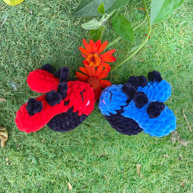 May include: Two handmade crochet ladybug toys, one red and one blue, with black accents and pom-poms. The toys are set on a bed of green grass, with orange flowers and green leaves in the background.