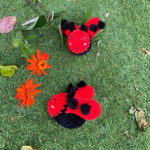 May include: Two handmade, plush ladybugs with red bodies, black spots, and black heads rest on green grass. Orange flowers and greenery are scattered around the ladybugs, creating a natural, whimsical scene.