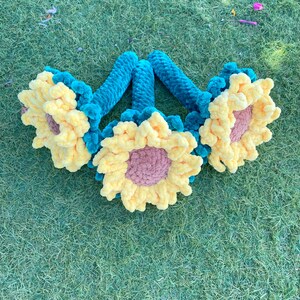 May include: Three crocheted sunflowers with teal stems. The sunflowers are yellow with a pink center.