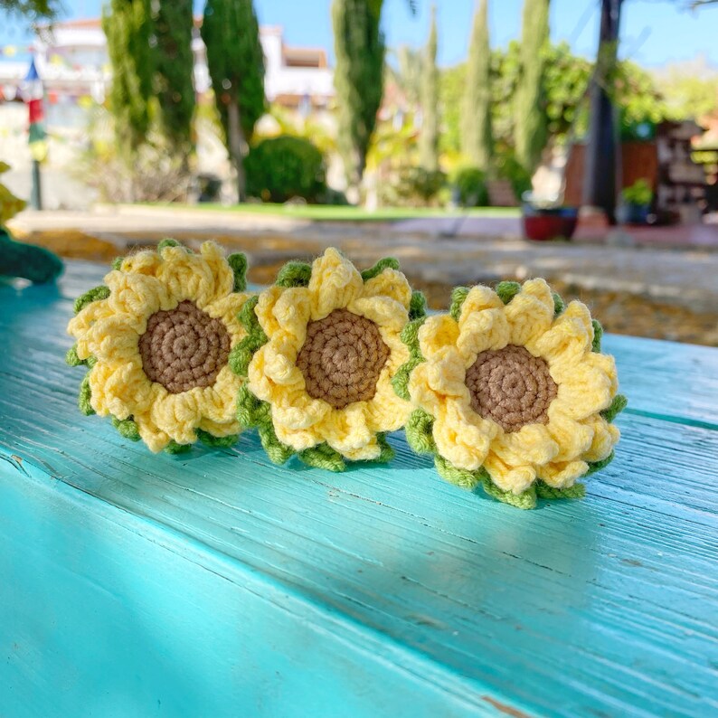 May include: Three crocheted sunflowers in yellow and green. The sunflowers are arranged in a row on a blue wooden surface.