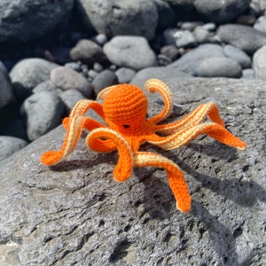 以下が含まれることがあります： 白いストライプのついたオレンジ色の編みぐるみタコ。タコはグレーのざらざらした表面の上に置かれています