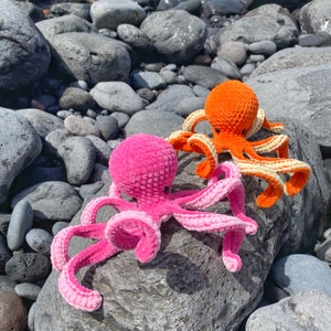 以下が含まれることがあります： ピンクとオレンジの2つの編みぐるみタコが、灰色の石のベッドの上に座っています。ピンクのタコは各触手に白いストライプがあり、オレンジのタコは各触手に黄色のストライプがあります。