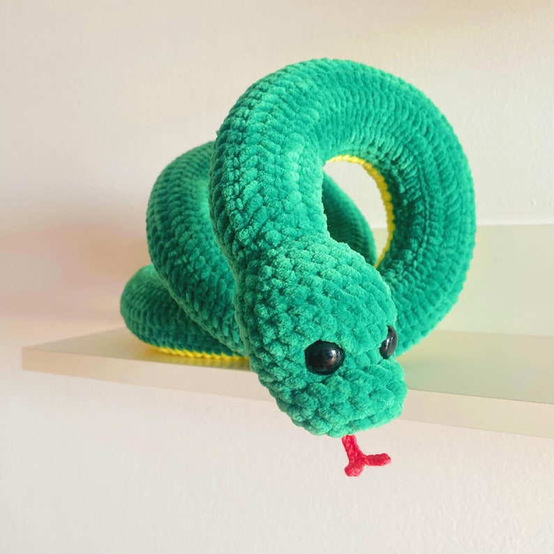 May include: A green crocheted snake plush toy with black eyes and a red tongue. The snake is curled up in a coil shape.