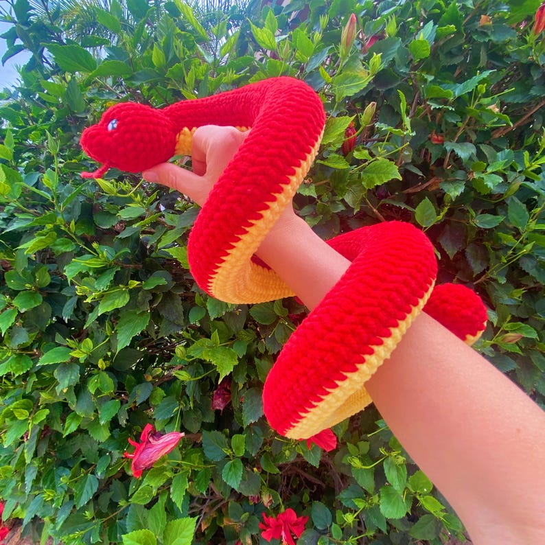 May include: A red and yellow crocheted snake toy. The snake is curled up and has a friendly face.